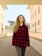 A woman with curly hair wearing a Red & Black Checkered SFP Flannel, made from a cotton-polyester blend, walks outdoors between buildings.