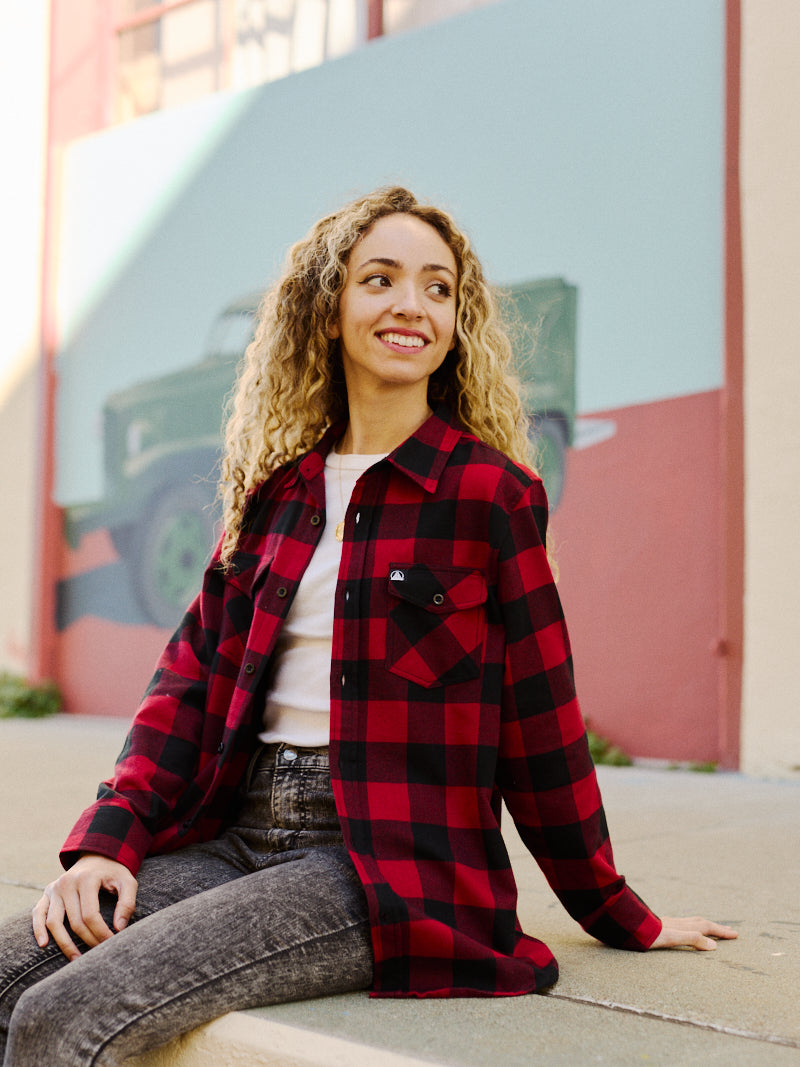 A smiling person with curly hair, wearing a Red & Black Checkered SFP Flannel that feels incredibly soft, sits in front of a colorful mural, their shirt enhancing the vibrant scene.