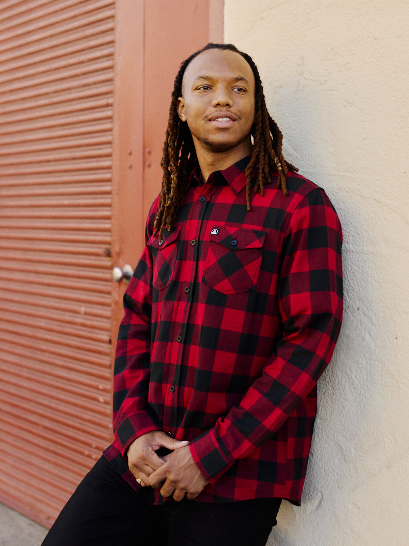 A person with long hair wearing a Red & Black Checkered SFP Flannel shirt stands against a wall, gazing to the side.