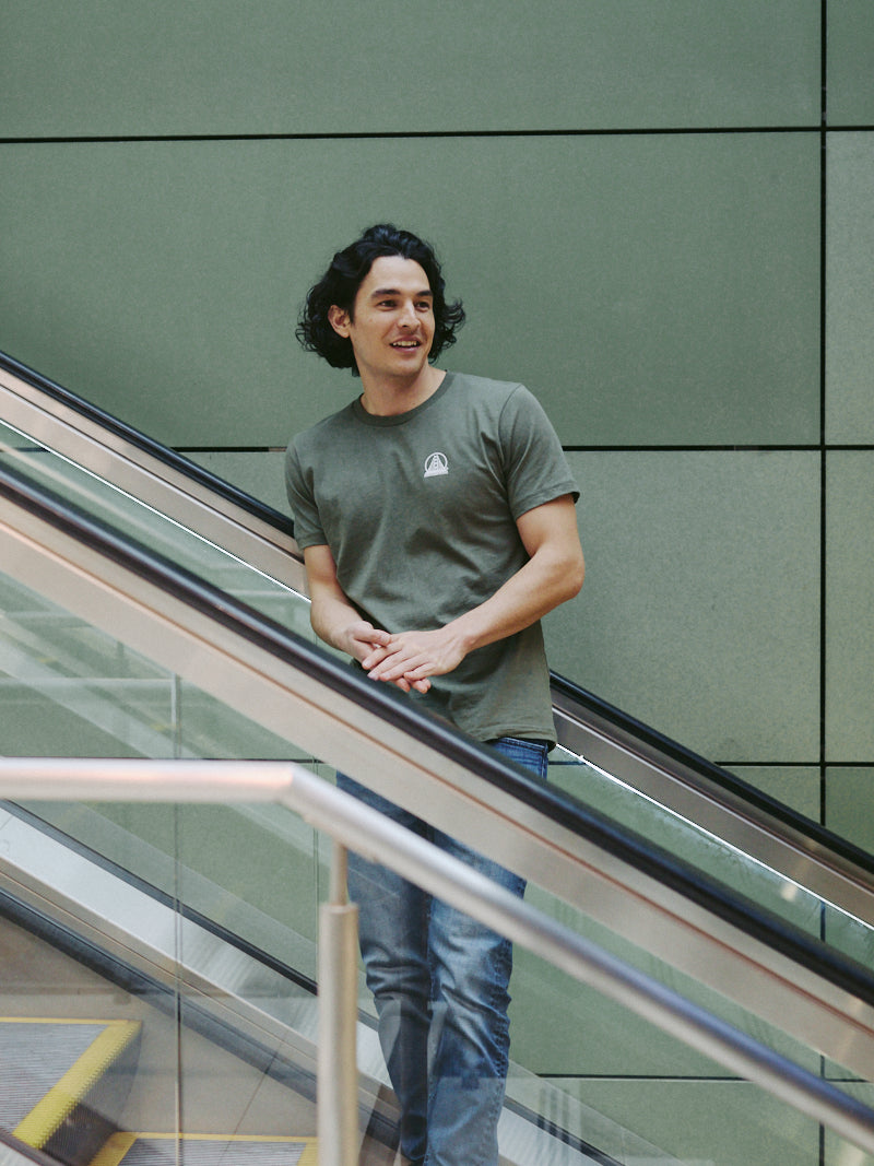 A person wearing a Military Green Mini Logo Tee and casual attire stands on an escalator against a green wall.