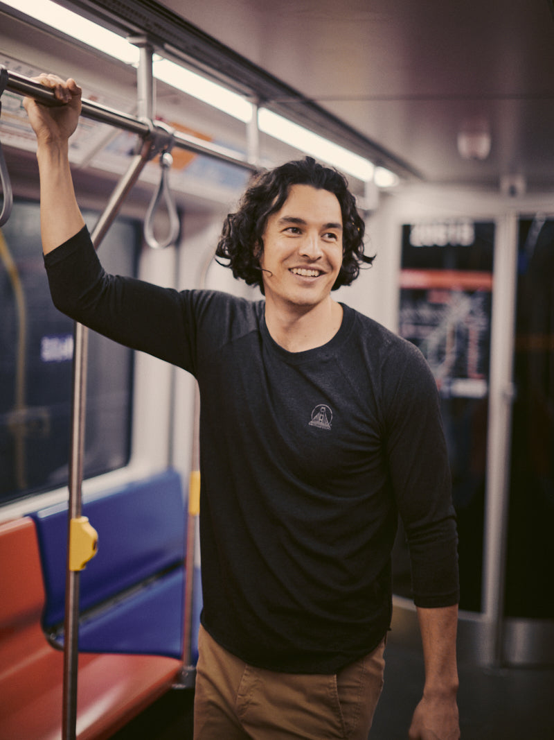 A man stands smiling inside a subway train, comfortably holding onto a pole. Dressed in everyday wear, his Charcoal Raglan w/ Mini Logo tee adds a touch of style to the journey. Empty seats scatter in the background as the train gently sways along its tracks.