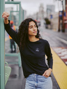 A person with long curly hair, dressed in a Charcoal Raglan w/ Mini Logo tee and jeans, leans against a structure at a train station, epitomizing the effortless style of everyday wear from our basics line.