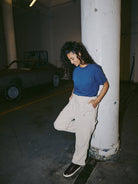 A person in a Heavyweight Midnight Pocket Tee and beige pants leans against a column in a dimly lit garage, with a vintage car adding to the ambiance.
