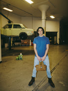 A person in a Heavyweight Midnight Pocket Tee holds a brown bag in a garage, with a car on a lift in the background.