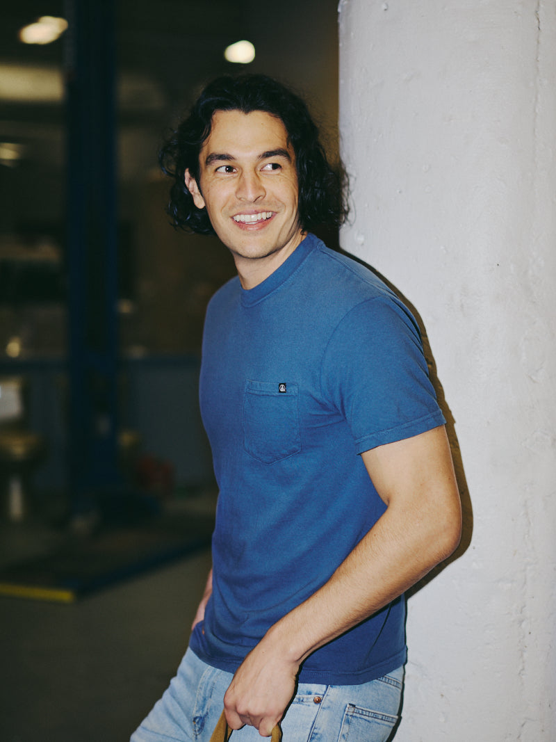 A smiling man in a blue shirt and jeans, reminiscent of the Heavyweight Midnight Pocket Tee, leans against a white pillar indoors.