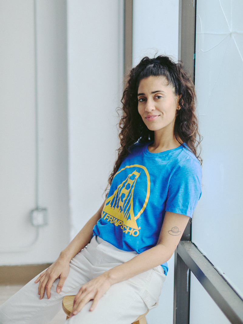 A person wearing our Women's Logo Tee in blue and gold from the basics line, adorned with a graphic design, sits on a stool by the window. Their long, curly hair frames their smiling face as they relish the comfort of the poly/cotton/rayon blend.