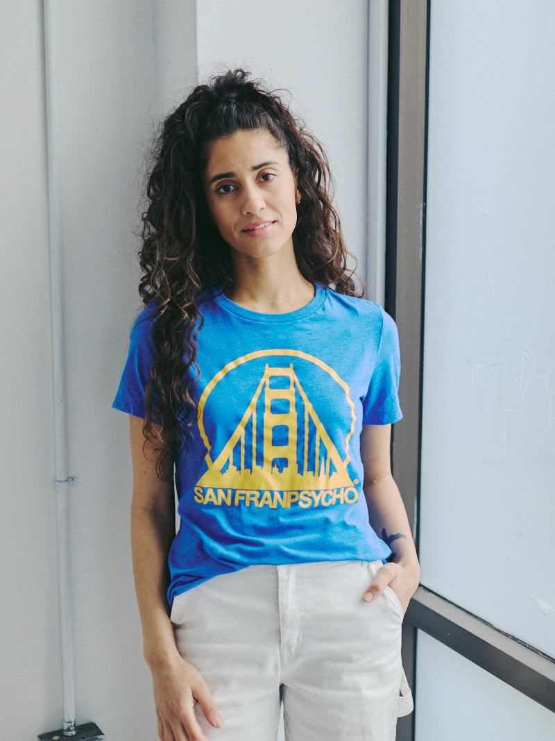 A person is standing by a window in a blue Women's Logo Tee - Blue & Gold, made from a comfy poly/cotton/rayon blend.
