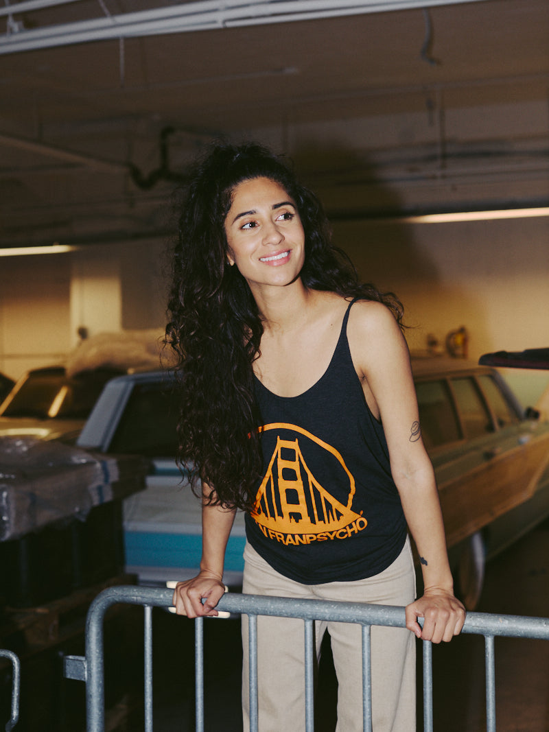 A smiling woman with curly hair, wearing the relaxed-fit Women's Logo Tank in charcoal and orange, leans on a metal railing in a parking garage.