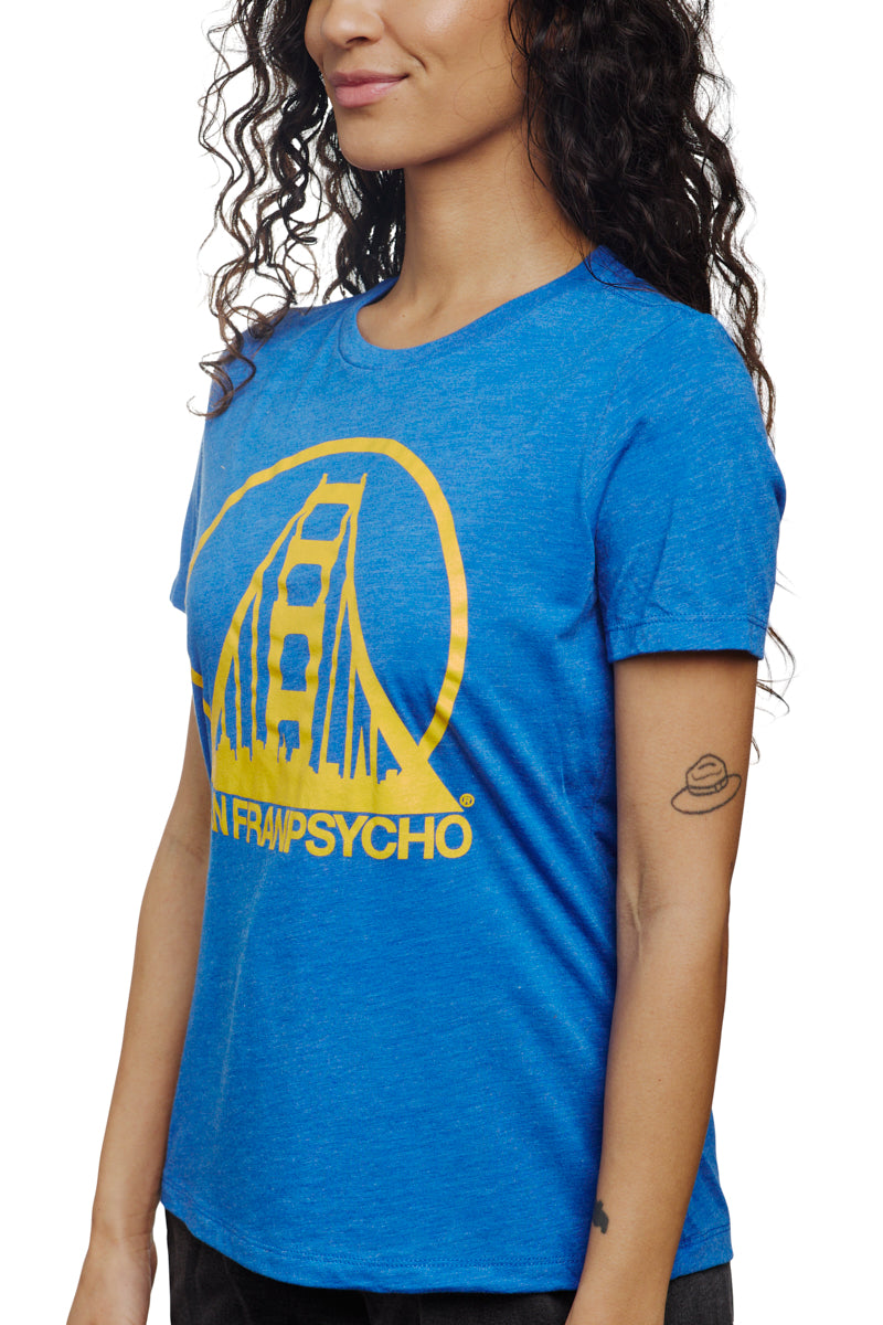 A woman wearing the Women's Logo Tee - Blue & Gold, designed from a blend of polyester, cotton, and rayon, smiles happily. The blue T-shirt displays a yellow bridge graphic alongside text. Her arm features a small planet tattoo, adding a unique touch to her casual style.