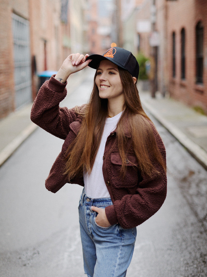 A woman wearing an Embroidered Logo Trucker Hat in black and orange smiles while standing on a brick-lined urban street.
