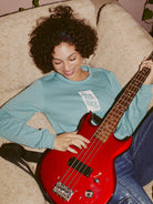 A person with curly hair, smiling as they play a red bass guitar, is lounging on a beige couch while wearing the Trippy Psycho Women's Crewneck from the Spring 2023 Collection, which is crafted from an ultra-soft cotton/polyester blend.