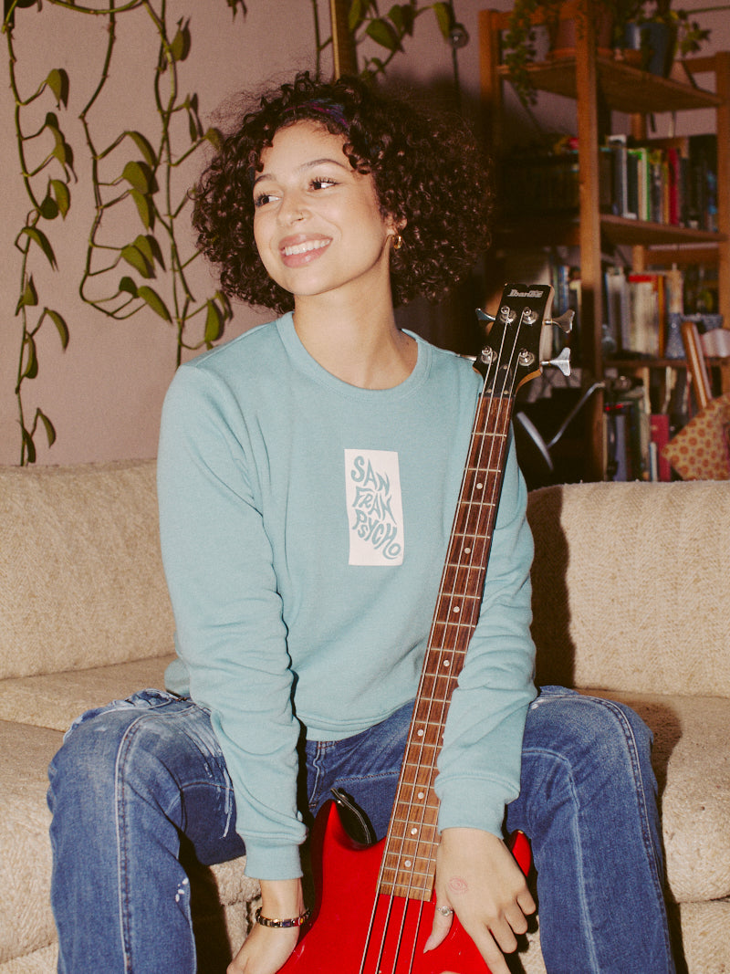 A person smiling, seated on a couch with a red bass guitar in hand, is wearing the Trippy Psycho Women's Crewneck. They're surrounded by plants and books, reveling in the comfort of its ultra-soft cotton/polyester blend fabric.