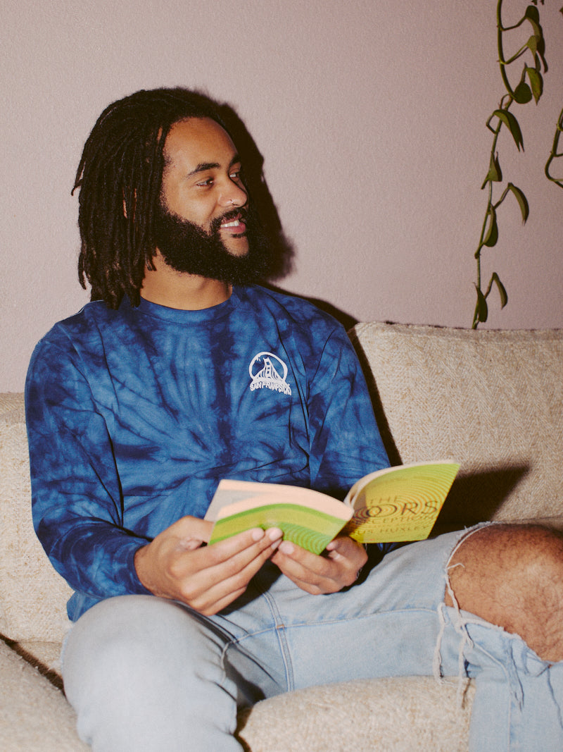 A man with dreadlocks reads a book while sitting on a beige couch, wearing the Tie-Dye Acid Logo Long Sleeve in navy and ripped jeans.