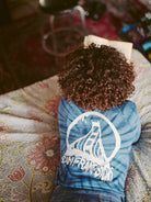 A person with curly hair lies on a bed reading, wearing a vibrant navy tie-dye long sleeve shirt featuring an acid logo.