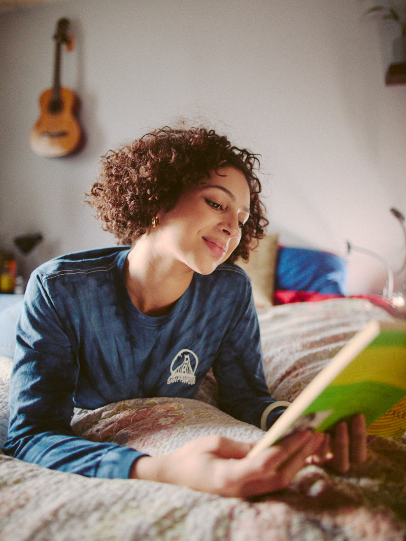 A person lounges on a bed, engrossed in a book, dressed in a cozy Tie-Dye Acid Logo Long Sleeve - Navy shirt. Nearby, a guitar hangs on the wall, adding to the room's relaxed and artistic vibe.
