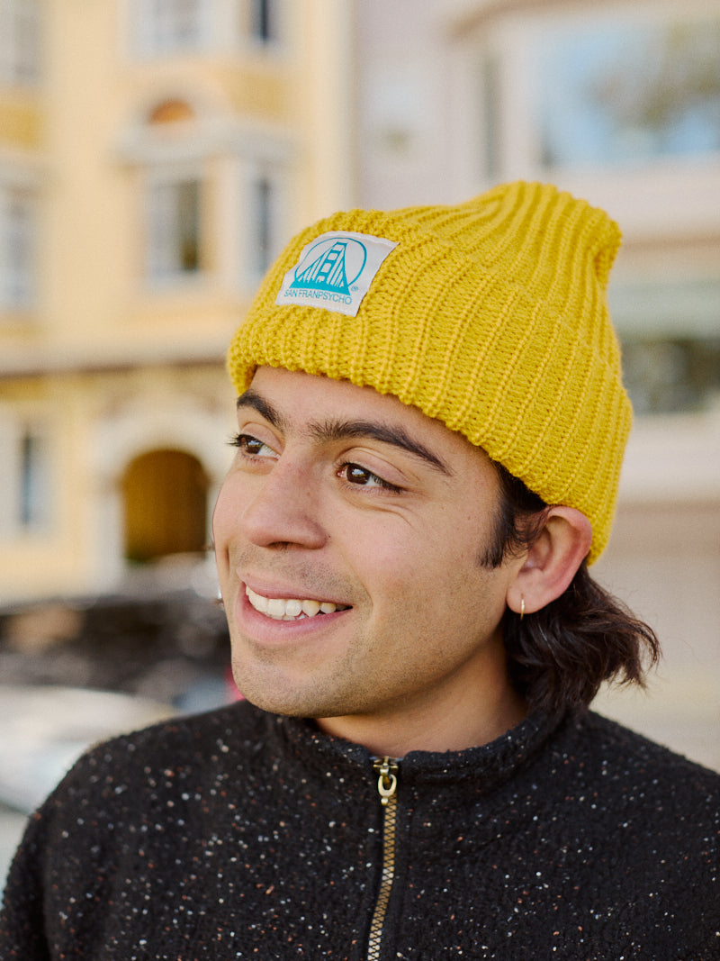 A person wearing a Mustard Lumberjack Beanie with a Natural/Turquoise SFP Logo and dark sweater smiles outside, against the backdrop of bustling San Franpsycho buildings.