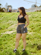 Dressed in Women's Embroidered Logo Jogger Shorts crafted from a soft cotton/polyester blend, a person in athletic wear stands on a grassy hill, smiling confidently with hands in pockets.