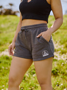 A person in a black tank top and the Women's Embroidered Logo Jogger Shorts stands on a lush grassy background. The gray shorts, crafted from a soft cotton/polyester blend, feature an embroidered bridge logo that adds a stylish touch to the casual ensemble.