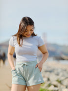 A person in a gray t-shirt and Women's Embroidered Logo Jogger Shorts made from a cotton/polyester blend stands on a rocky beach, looking down and smiling.