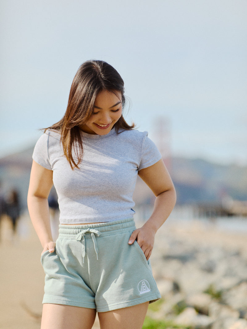 A person in a gray t-shirt and Women's Embroidered Logo Jogger Shorts made from a cotton/polyester blend stands on a rocky beach, looking down and smiling.