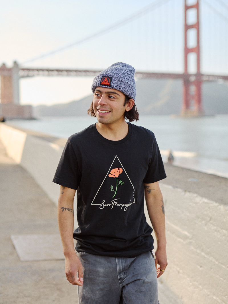 A person in a beanie and the black California Poppy Tee smiles near a waterfront, with a large bridge in the background.