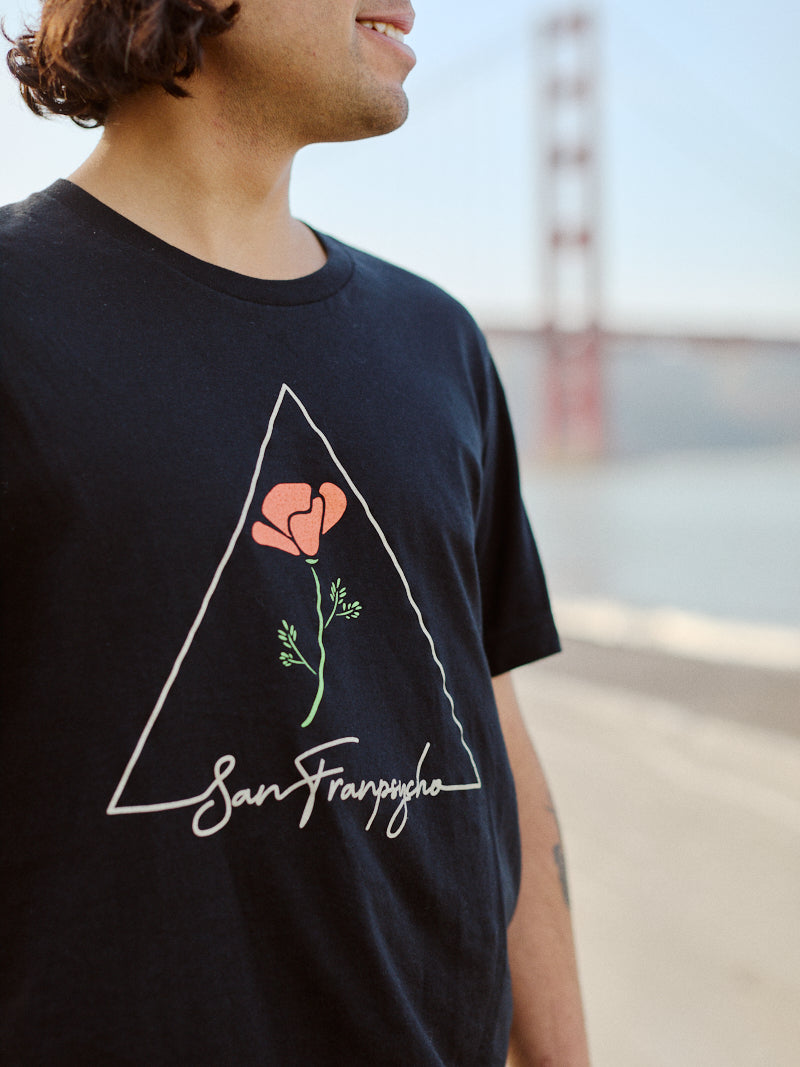 A person wearing the California Poppy Tee, a 100% cotton unisex shirt featuring a black poppy design, stands near a blurred view of the Golden Gate Bridge.