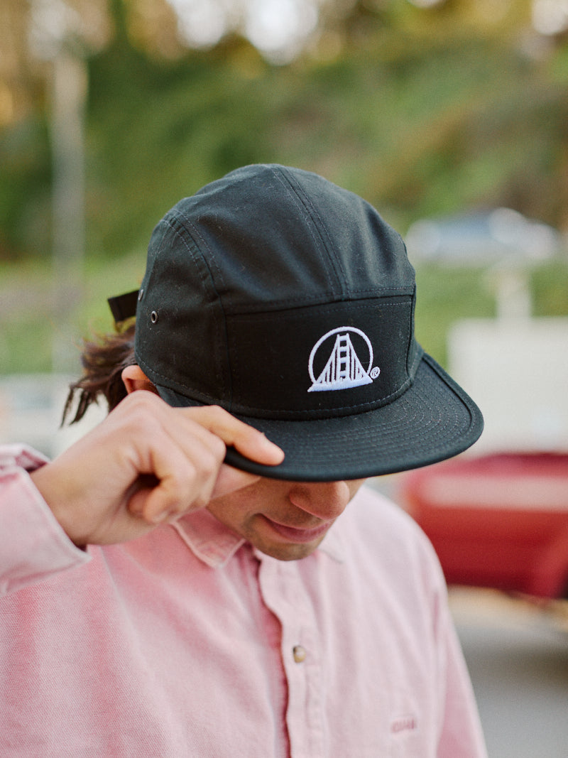 The individual in a pink shirt confidently adjusts the brim of their black Logo Camper Hat adorned with an SFP logo.