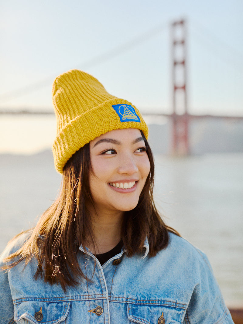 A cheerful person wearing a mustard lumberjack beanie with the blue/yellow SFP logo and a denim jacket stands near a large red bridge over water, embodying the quintessential San Franpsycho vibe.