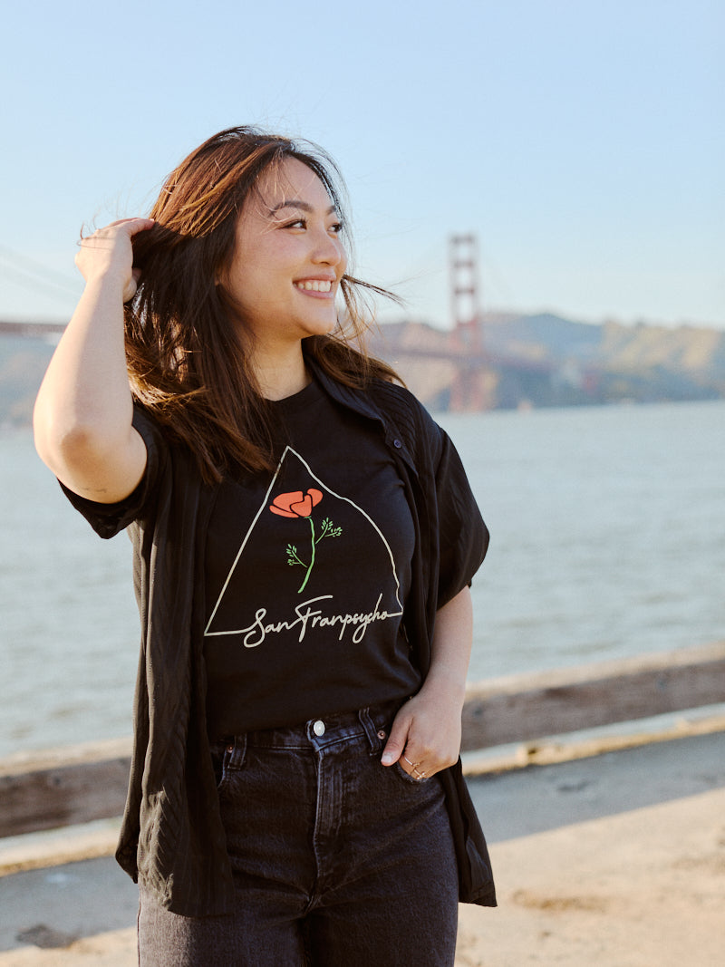A smiling woman stands by the water with the Golden Gate Bridge in the background, wearing a California Poppy Tee from Black Poppy. This 100% cotton unisex shirt perfectly captures the spirit of San Francisco.