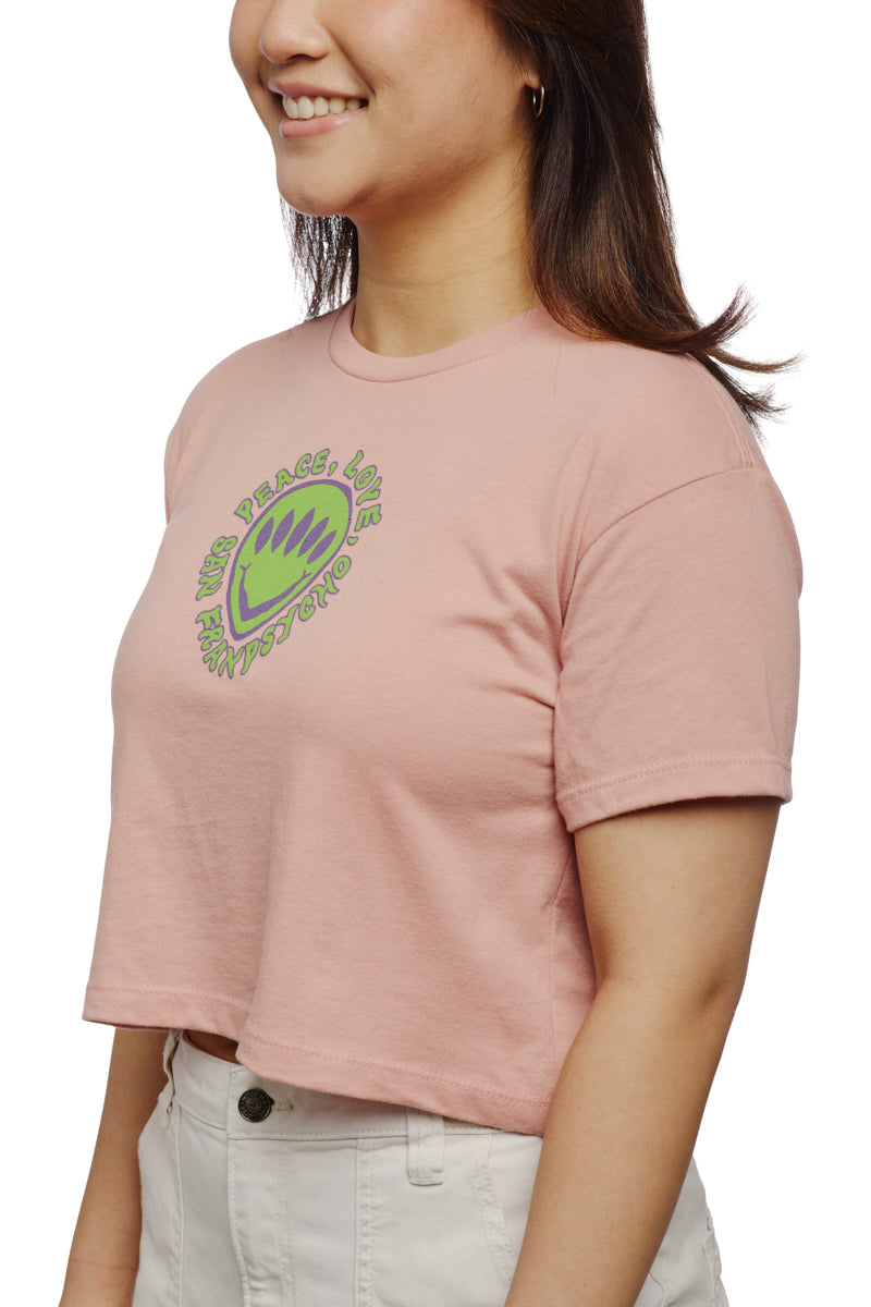 A smiling person wears a pink Peace Love Alien Crop Top paired with white pants, radiating joy and harmony.
