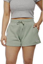 A person wearing a gray cropped top and Women's Embroidered Logo Jogger Shorts in light green, crafted from a comfortable cotton/polyester blend, casually rests one hand in the pocket.