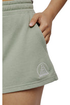 Close-up of a person wearing Women's Embroidered Logo Jogger Shorts in light green, made from a cotton/polyester blend, featuring a pocket and an embroidered bridge design on the hem.