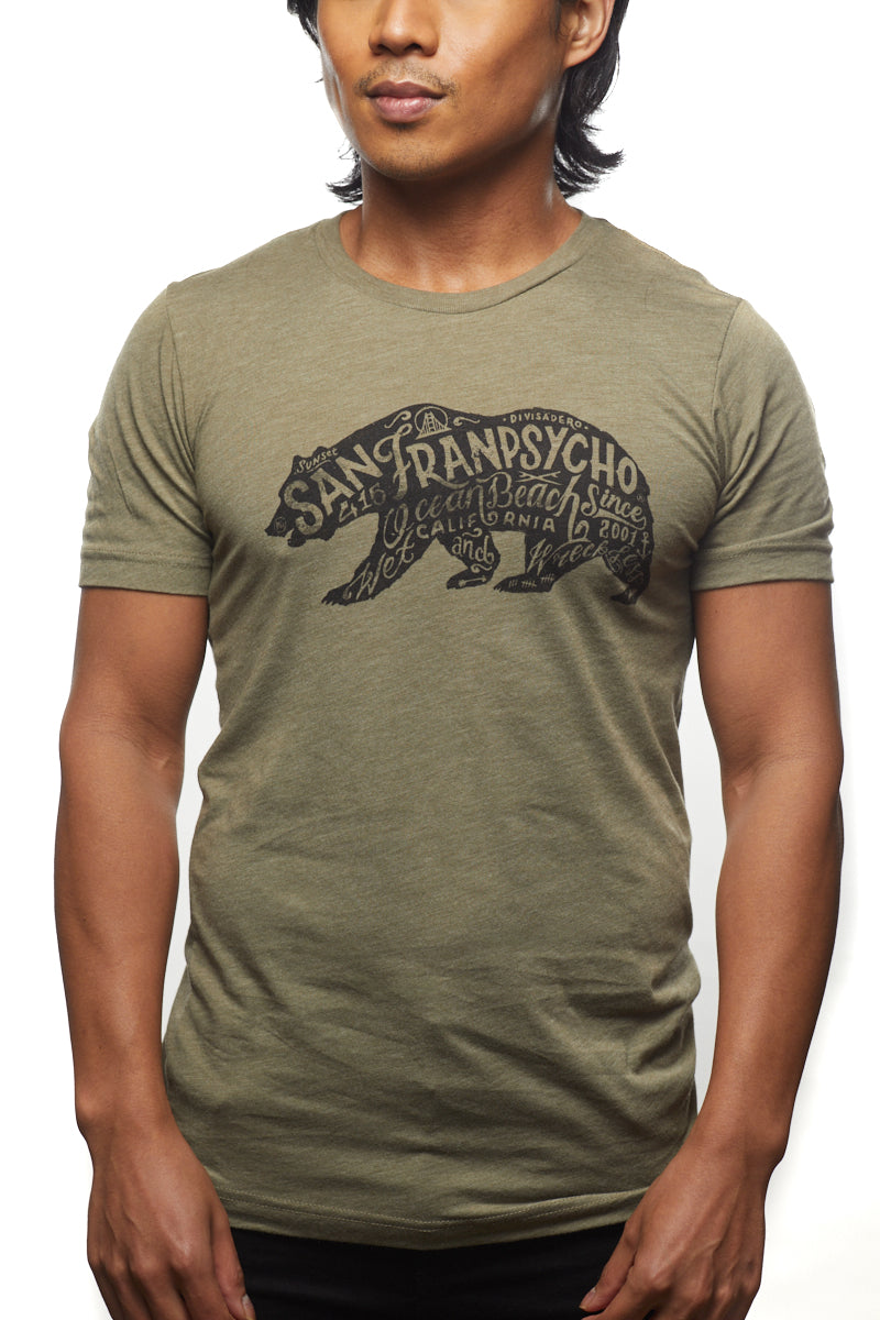 An individual wearing the Cali Bear Tee in olive green stands out in the Sunset District, showcasing a striking design with bold black text.