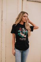 A person in an incredibly soft black Surf Club Tee stands against a light wall, gently touching their hair.