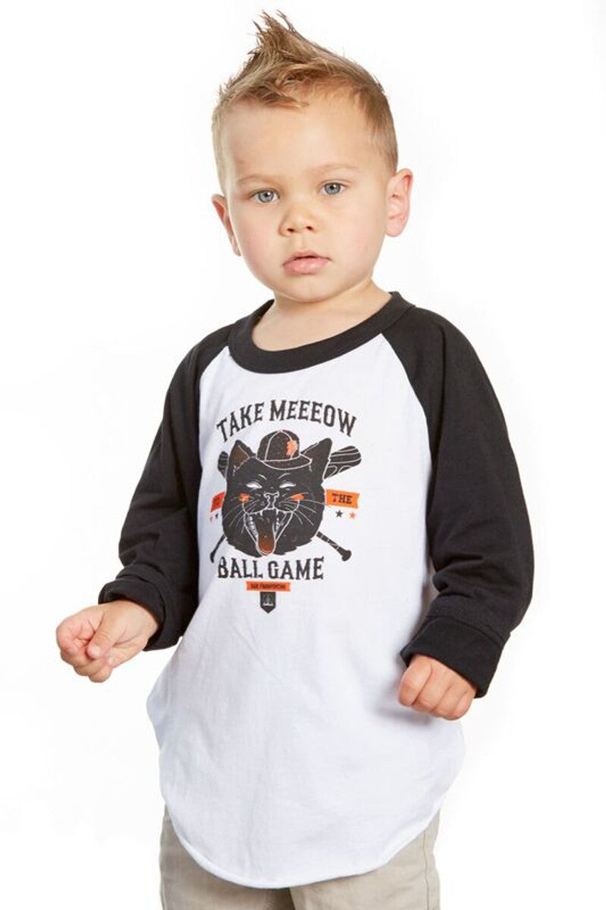 A young child in a white and black Youth Take Meeeow Baseball Tee, featuring a playful cat graphic and "Take Meow to the Ball Game" text, is perfect for celebrating baseball season in San Francisco. Available in kids' sizes.