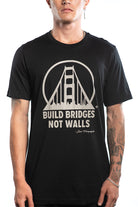 A person wearing the Build Bridges Not Walls Tee, a hand-printed black shirt featuring a bridge graphic and the text "Build Bridges Not Walls.