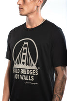A person wearing the Build Bridges Not Walls Tee showcases a hand-printed black shirt featuring a bridge graphic and the inspiring message "Build Bridges Not Walls" prominently displayed.
