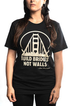 A person is wearing the "Build Bridges Not Walls Tee," a hand-printed black shirt showcasing a bridge graphic with the bold slogan, "Build Bridges Not Walls.