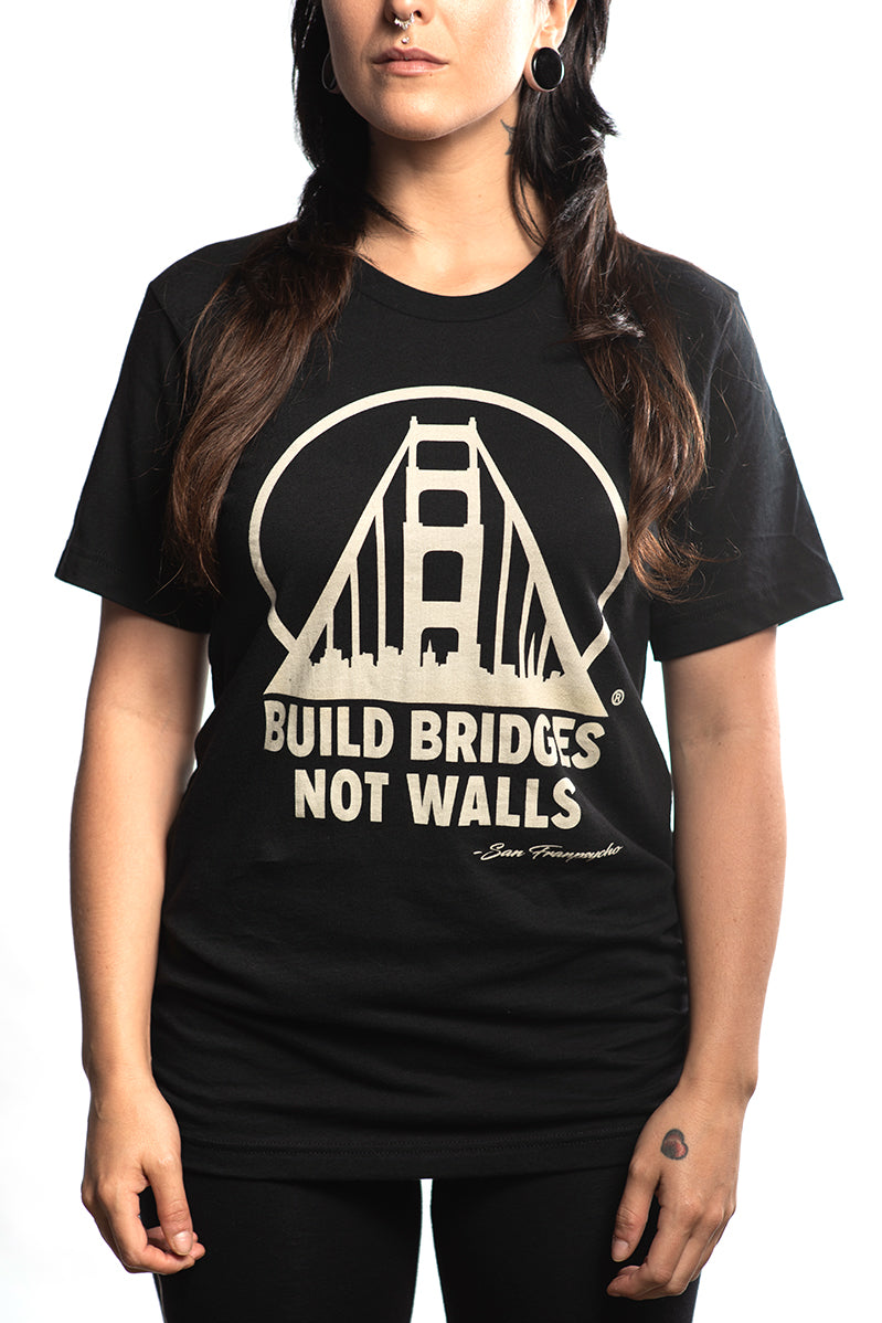 A person is wearing the "Build Bridges Not Walls Tee," a hand-printed black shirt showcasing a bridge graphic with the bold slogan, "Build Bridges Not Walls.