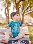 A baby sits on a picnic blanket outdoors, dressed in a Teal Fogtown Onesie. Surrounded by trees, the onesie, made of 100% cotton, adds to the perfect day in nature.