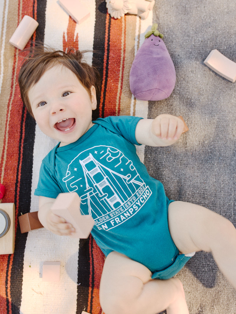 The baby, dressed in a Teal Fogtown Onesie made from 100% cotton, is surrounded by toys while lying on a striped blanket, smiling brightly as they hold blocks.