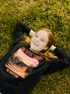 A girl lies on the grass, smiling, in her Youth Outer Sunset Crewneck featuring a hand-printed, colorful graphic of San Francisco's Outer Sunset.