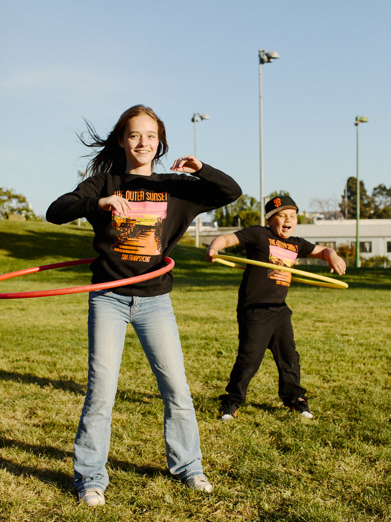 Two kids are hula hooping on the grass in San Francisco's Outer Sunset, both wearing Youth Outer Sunset Crewnecks featuring hand-printed orange designs, under a clear blue sky.