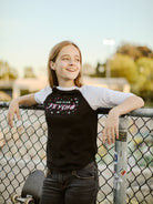 A girl in a Youth Planet Rock Baseball Tee smiles while leaning on a fence, her skateboard resting nearby, exuding 90s vibes with the sprawling park in the background.