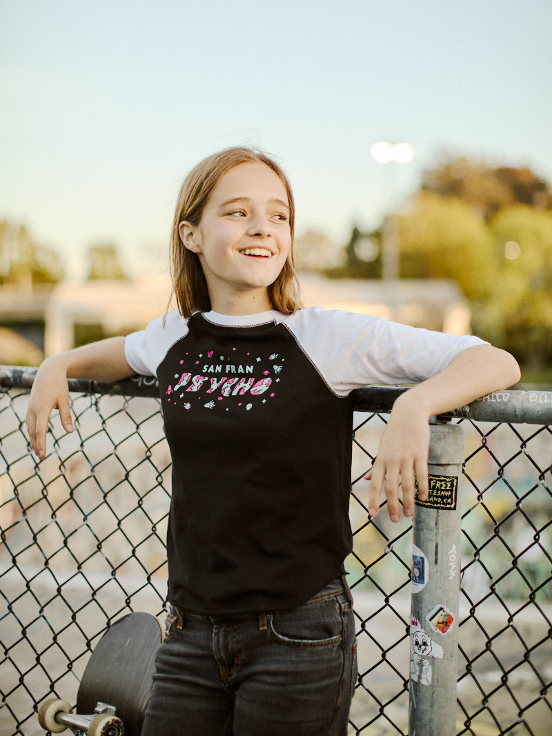 A girl in a Youth Planet Rock Baseball Tee smiles while leaning on a fence, her skateboard resting nearby, exuding 90s vibes with the sprawling park in the background.