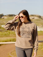 A woman wearing sunglasses and a Desert Daze Crewneck from the Summer 2023 line stands outside on a bright day, highlighting its unisex design.