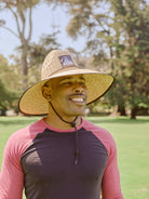 A smiling man stands outdoors in a park on a sunny day, enjoying sun protection with the Lifeguard Hat.