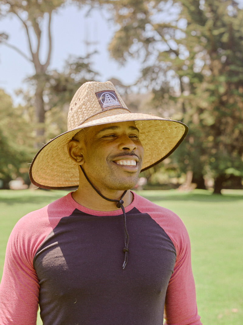A smiling man stands outdoors in a park on a sunny day, enjoying sun protection with the Lifeguard Hat.