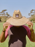 Wearing the Lifeguard Hat, a person stands on grass with trees and a blue sky behind them, holding the hat’s brim for stylish sun protection.