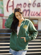 A woman in a Hippie Hill Zip-Up Hoodie smiles, standing outdoors with a blurred music sign in the background, capturing the lively vibes of Hippie Hill.